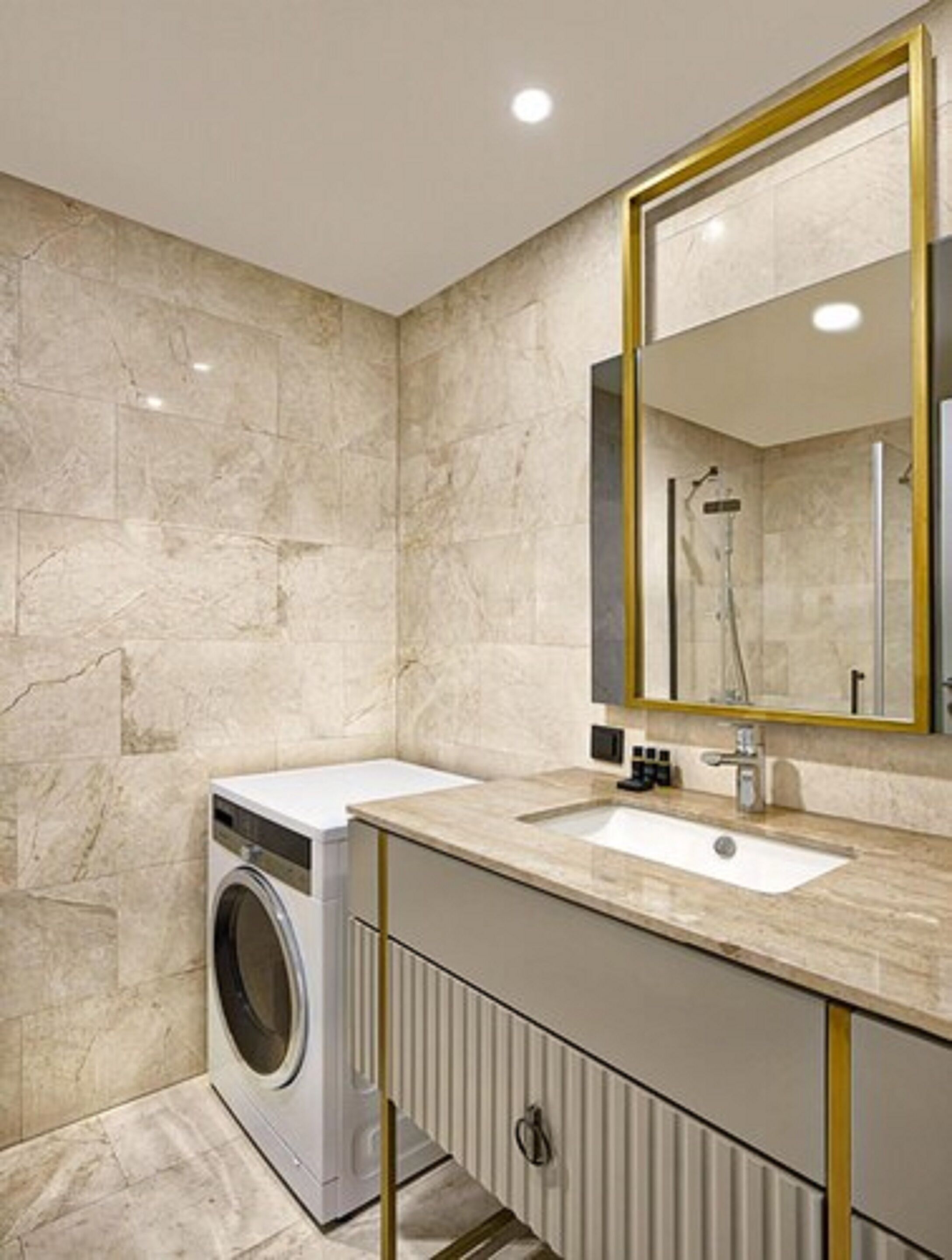 A modern bathroom with a marble wall, a washing machine, a sink with a gold-framed mirror, and a shower.