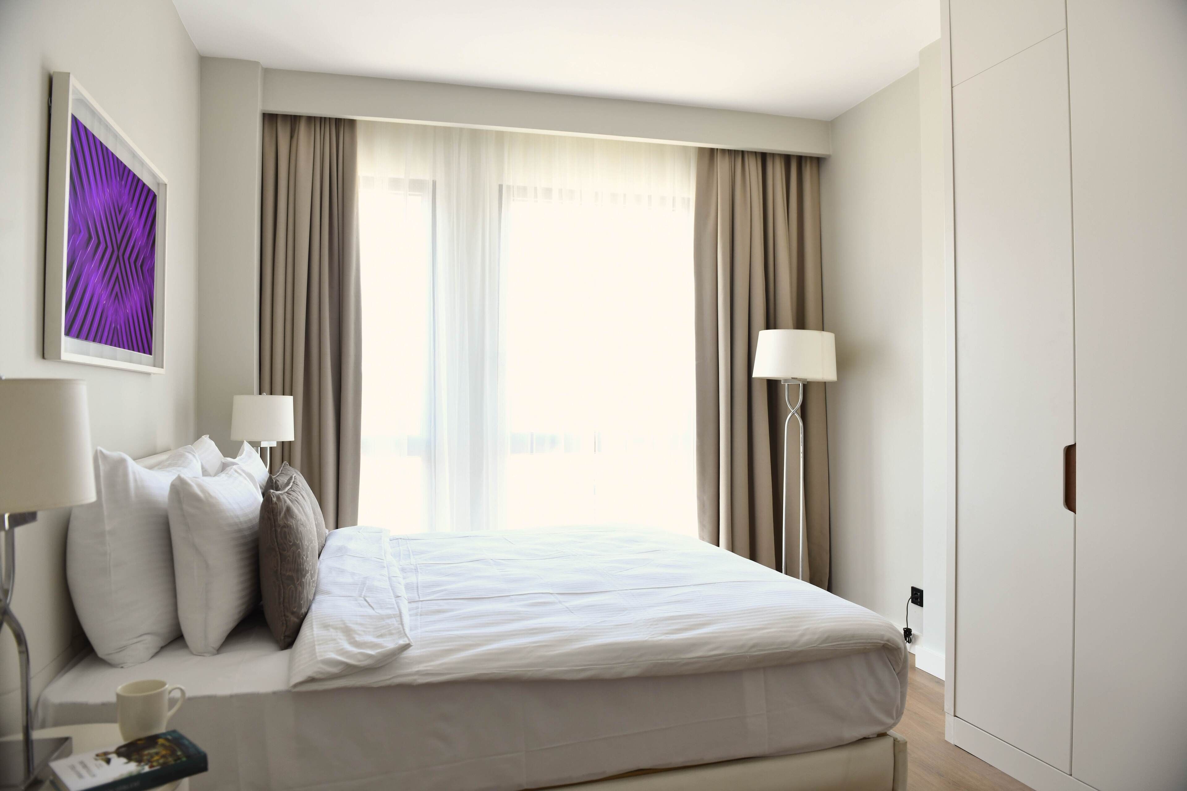 A hotel room with a bed, a bedside table with a lamp, a coffee cup, and a framed picture on the wall.