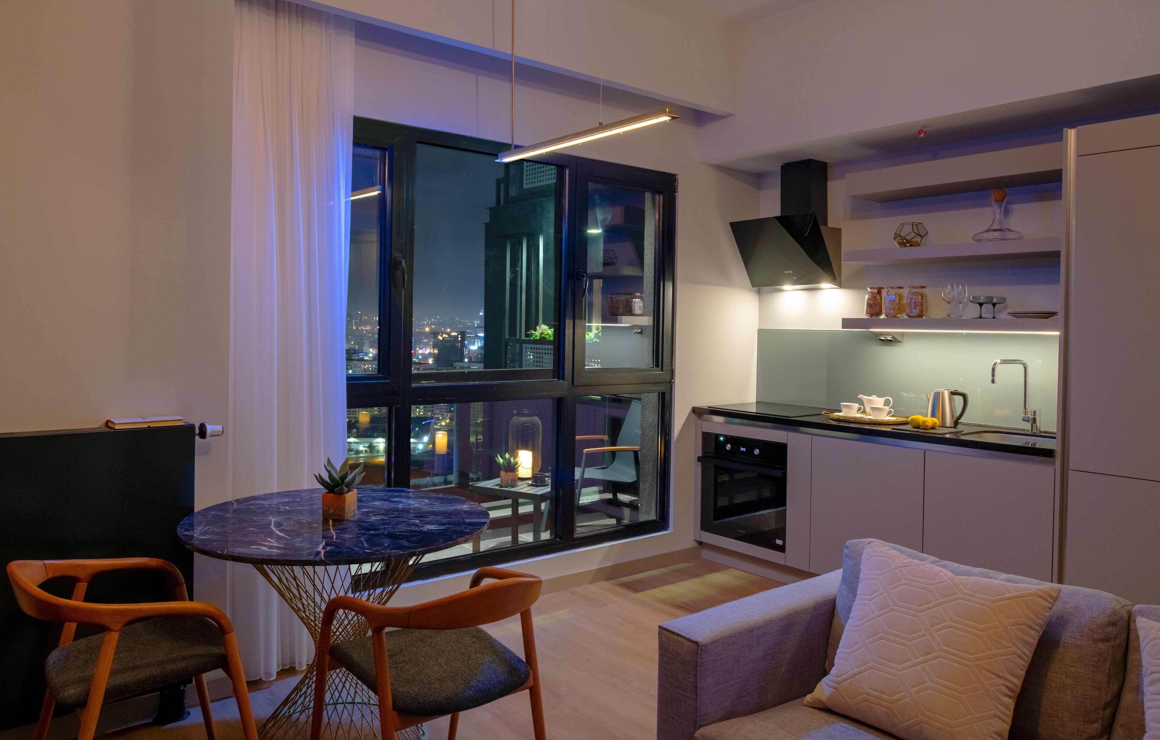 A modern kitchen with a large window, a dining area with a round table, and a sofa with a cushion.