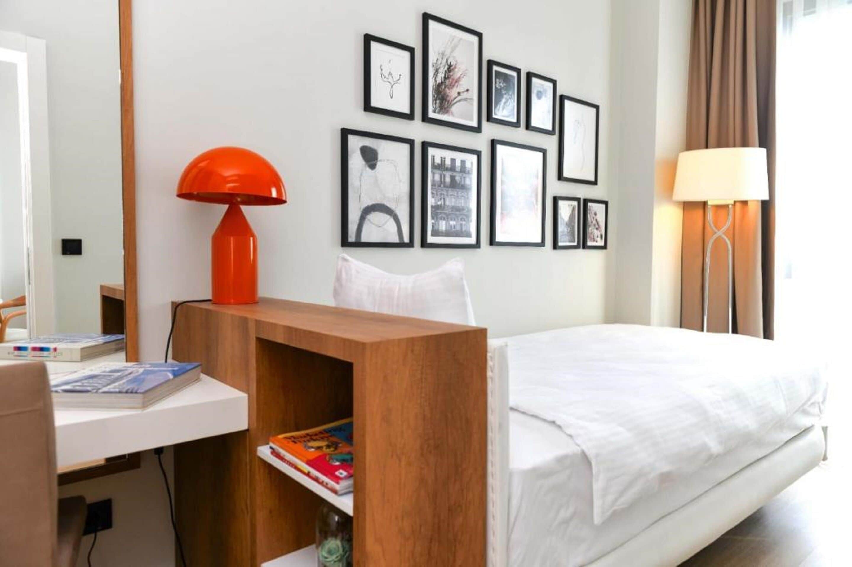 A hotel room with a bed, a desk with a chair, a wooden side table, a red lamp, a white lamp, and a wall with framed pictures.