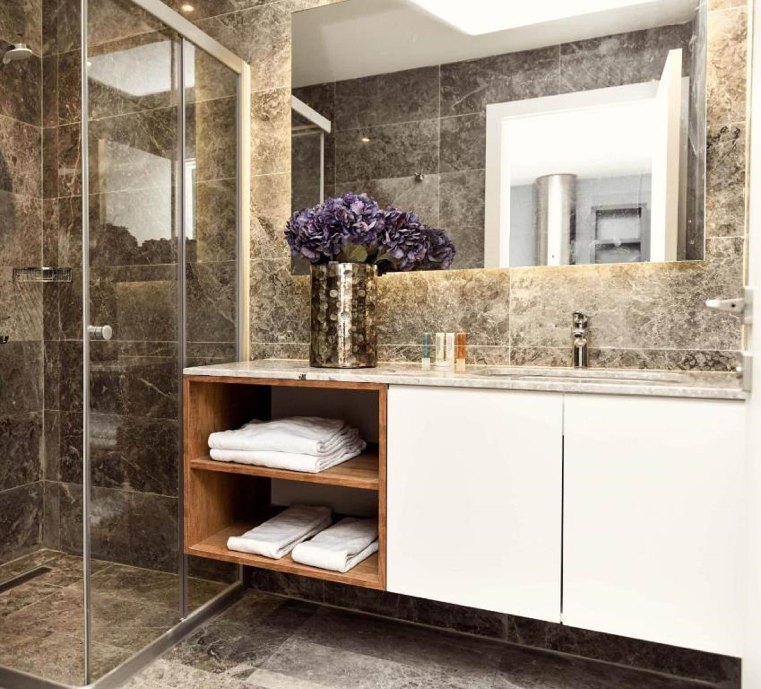A modern bathroom with a glass shower enclosure, marble tiles, a vanity with a mirror, and a shelf with folded towels.