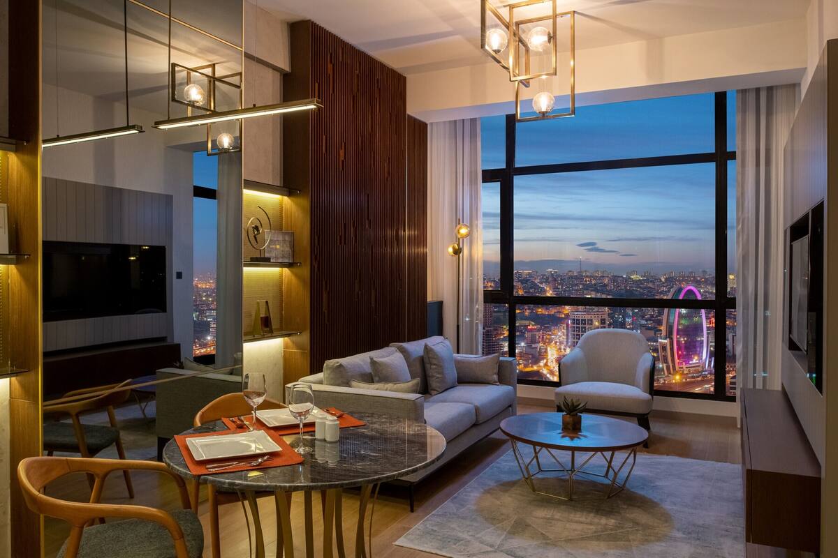 A modern living room with a city view, a sofa, a coffee table, and a dining area with chairs and a table set for two.