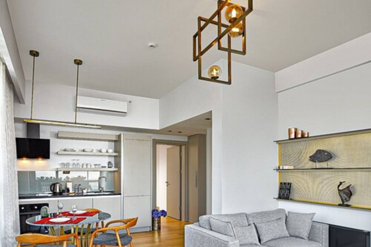 A modern living room with a grey sofa, wooden dining table, and chairs. There is a kitchen in the background with white cabinets and a TV mounted above the counter.
