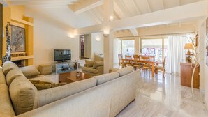 A spacious living room with a large sofa, a television, and a dining area with wooden furniture. - Villa Anna Maria 3 Bedroom Private Pool (Heraklion)