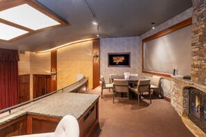 Un intérieur chaleureux avec une cheminée en pierre, un coin repas avec des chaises, un bar avec un comptoir et un panneau indiquant « soirée cinéma ».