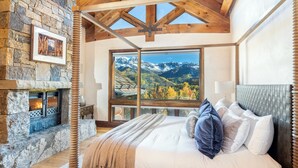 A bedroom with a stone fireplace, a large window with a mountain view, a bed with pillows, and wooden beams on the ceiling. - PICTURE PERFECT- Stunning Ski-In/Out Trailside Manor Perfect for Groups (Mountain Village)