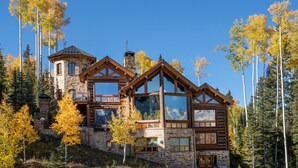 A log cabin with multiple windows and a stone chimney, surrounded by tall trees and a clear sky. - PICTURE PERFECT- Stunning Ski-In/Out Trailside Manor Perfect for Groups (Mountain Village)
