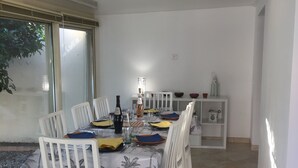 Un coin repas avec une longue table dressée pour six personnes, des chaises blanches, un petit meuble blanc et une grande fenêtre donnant sur un paysage verdoyant.