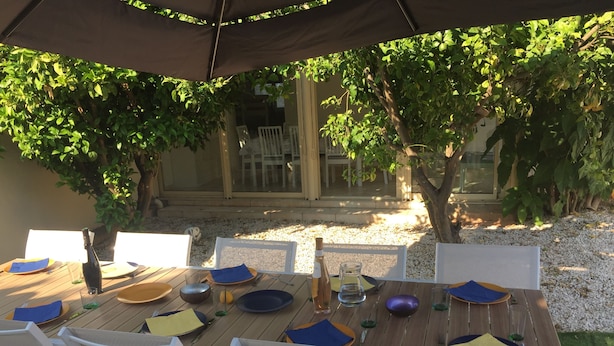 Un espace de restauration en plein air avec une table en bois et des chaises blanches, entouré de verdure et doté d’un patio couvert.