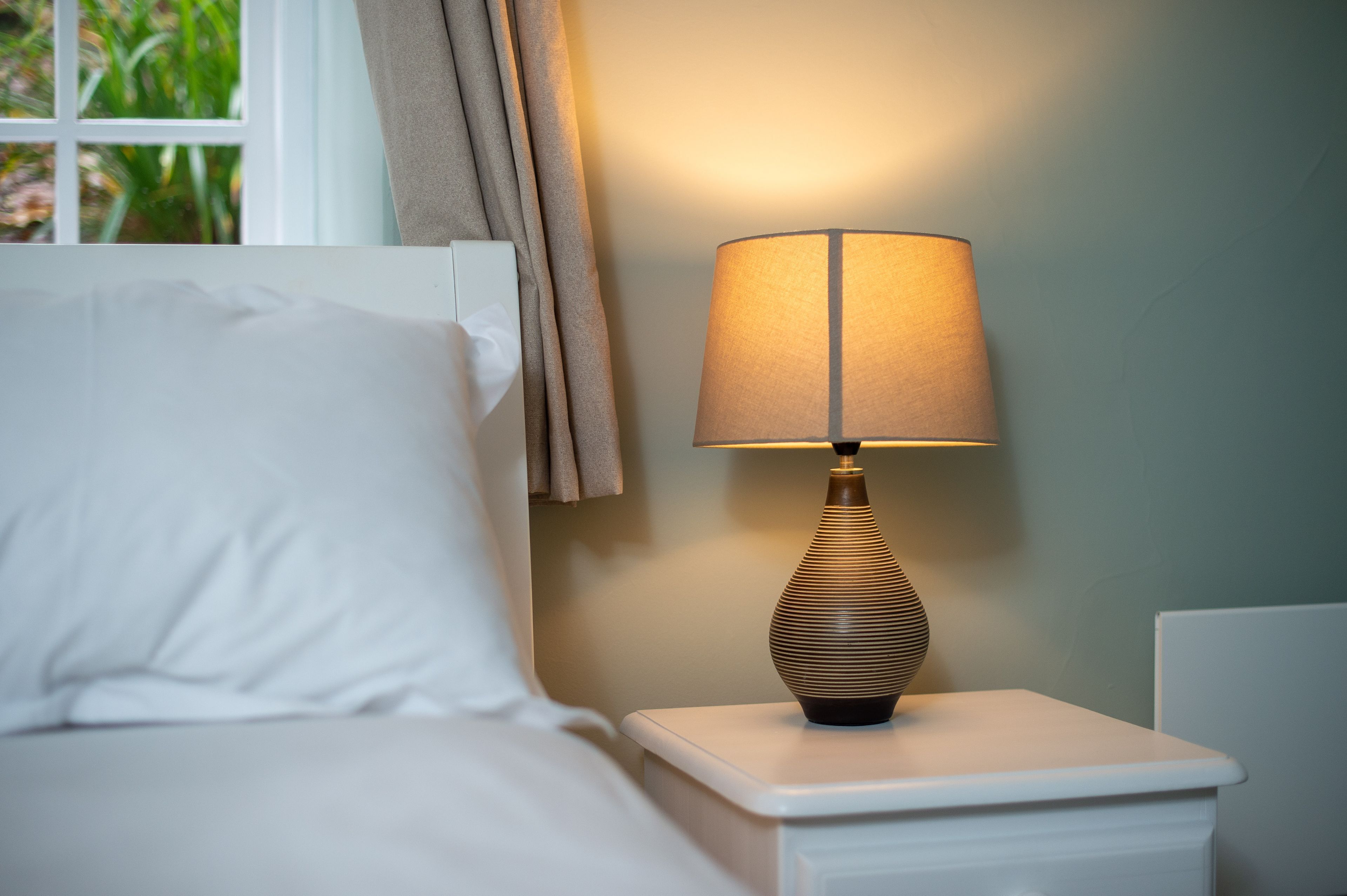 A neatly made bed with white linens, a bedside lamp with a ribbed base, and a window with curtains.