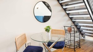 Habitació moderna amb taula de vidre, dues cadires, un mirall, una petita planta i una taula auxiliar de metall.
