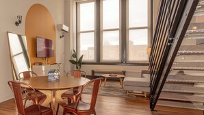 Et moderne spiseområde med et rundt bord, kobberfarvede stole, et fjernsyn og et stort vindue med udsigt til bygninger.
