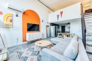 A modern living room with a grey sofa, a wooden coffee table, and a TV mounted on an orange wall. There is a dining area with a wooden table and chairs, and a staircase leading to an upper level.