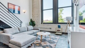 A modern living room with a grey sectional sofa, a round coffee table, and a large window with a view of buildings and trees. - Sosuite | Studio Loft w W/D, Gym, Lounge (Philadelphia)