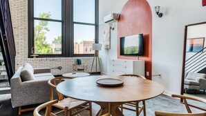A modern interior with a round wooden table, a grey sofa, and a large window offering a view of buildings outside. - Sosuite | Studio Loft w W/D, Gym, Lounge (Philadelphia)