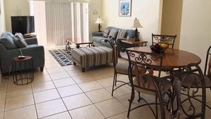 A living room with a sofa, a dining table with chairs, a TV, and a window with blinds. - Beautiful Golf View Southern Dunes 4 Bedroom Villa by RedAwning (Haines City)