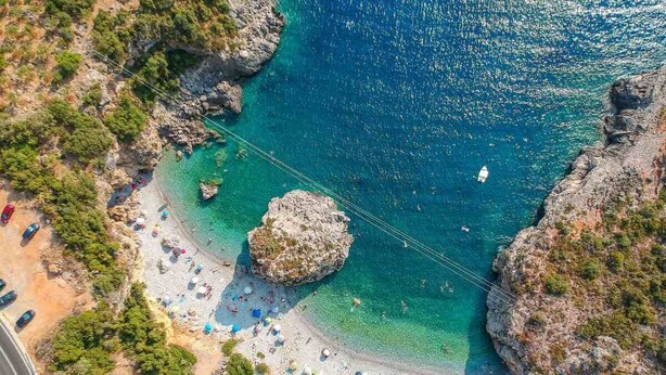 Vista aérea de una playa costera con aguas turquesas cristalinas, formaciones rocosas y una playa de arena con sombrillas y gente.