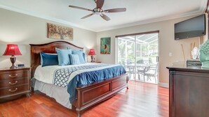 A bedroom with a wooden bed, two bedside tables with lamps, a ceiling fan, a TV mounted on the wall, and a sliding glass door leading to an outdoor area.