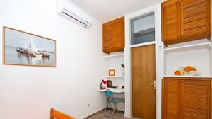 Habitación pequeña con cama, escritorio y silla. Se observa un armario de madera y una estantería con toallas.