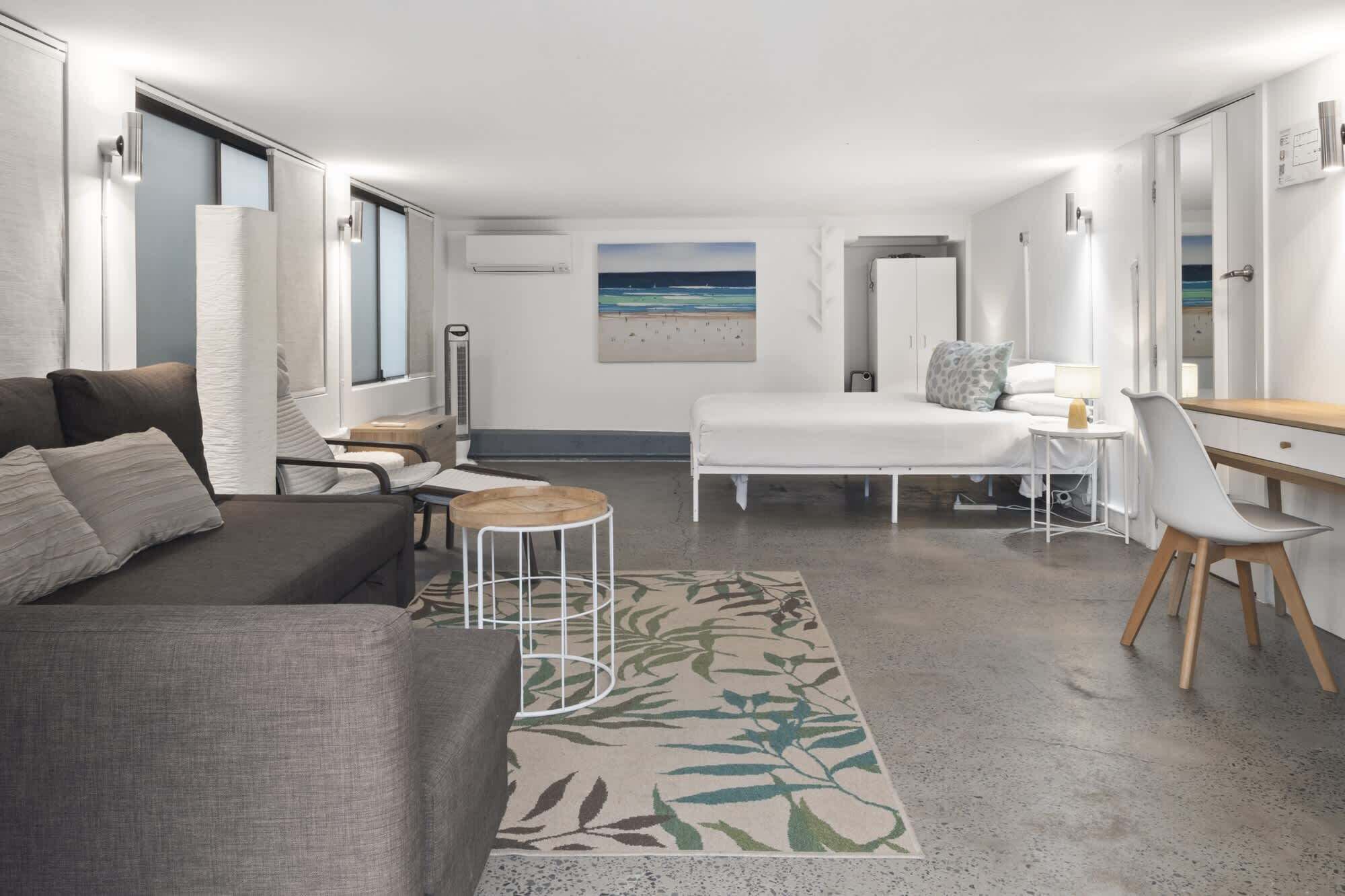 A modern hotel room with a bed, a desk, a chair, a sofa, and a large window with a view of the beach.