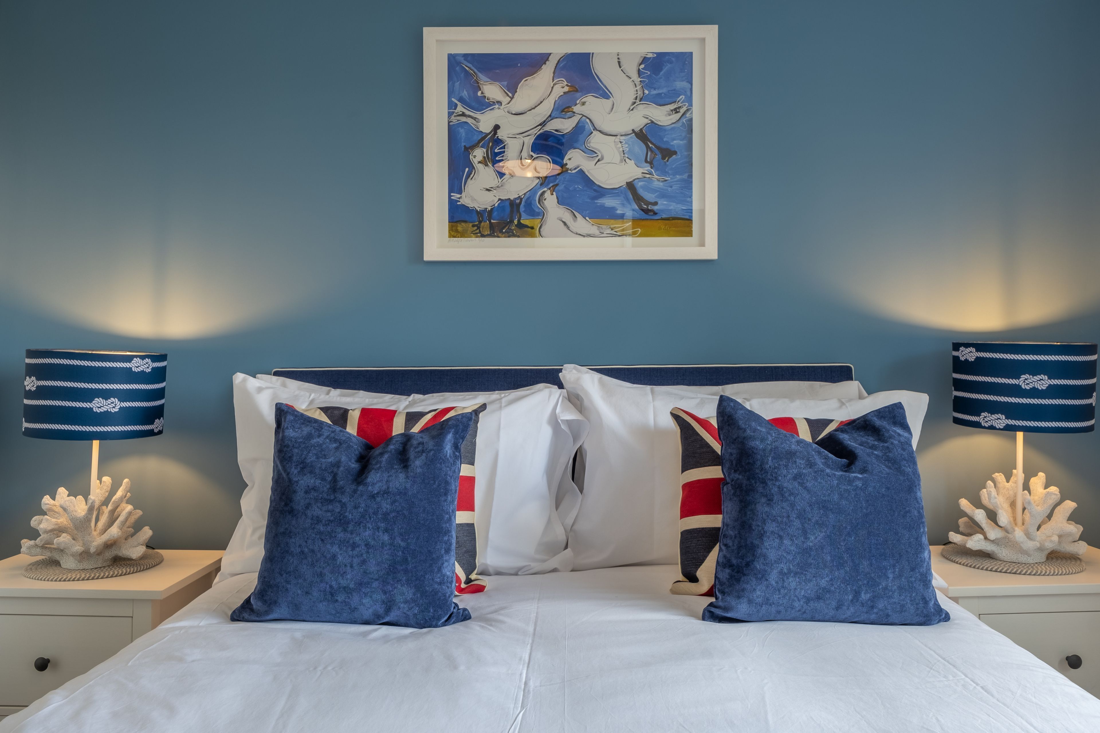 A neatly made bed with white linens and blue pillows, flanked by two nightstands with decorative corals and striped lamps.