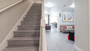 Uma escadaria leva a uma sala de estar com um sofá cinzento, uma almofada vermelha e branca, e quadros nas paredes.