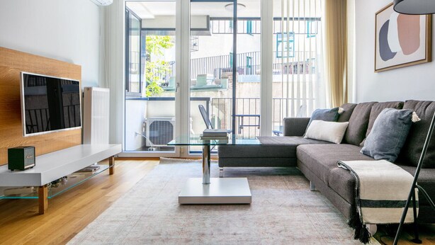 Un salon moderne doté d’un téléviseur à écran plat, d’une table basse en verre, d’un canapé et offrant une vue sur un balcon aménagé avec du mobilier d’extérieur.