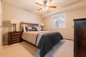 Une chambre à coucher comprenant un lit, des tables de chevet, un ventilateur de plafond, une fenêtre et une commode en bois foncé.