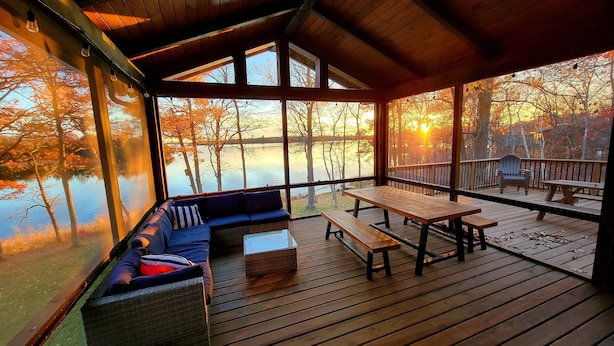 A wooden deck with a sofa and a table, overlooking a lake with autumn trees.
