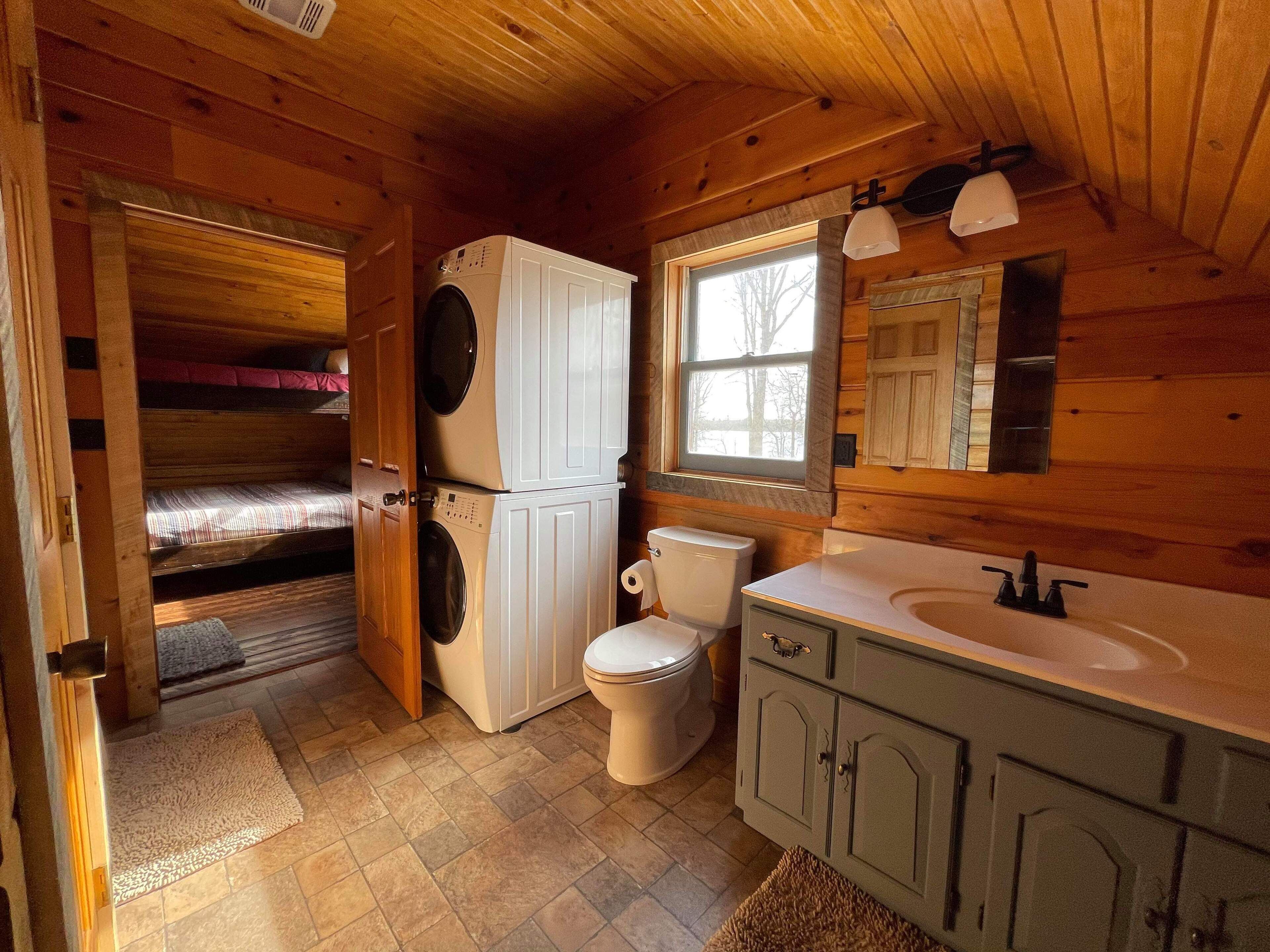 Un bagno in una cabina di legno compatta, con lavatrice, toilette, lavandino e una finestra che lascia entrare luce naturale.