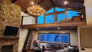 A spacious loft-style living area with a stone fireplace, a wooden staircase, a large chandelier, and a view of the sky through multiple windows.