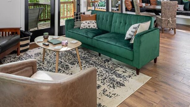 Amplio salón con un sofá verde, una mesa redonda de centro y suelo de madera. Hay ventanas grandes y una escalera de madera.