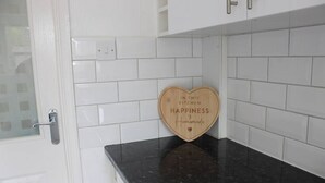 Uma cozinha com paredes revestidas de azulejos brancos, bancada preta e um letreiro em formato de coração de madeira com a inscrição 'FELICIDADE é feita em casa'.