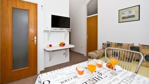 A small room with a wooden door, a TV on a white shelf, a table with breakfast items, and a sofa with cushions. - A3 - Best Location in Center With the sea View (Vodice)
