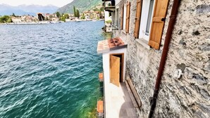 A narrow stone building with a wooden door and window shutters, overlooking a calm lake with buildings in the background. - VILLA MARIA by Design Studio (Bellano)