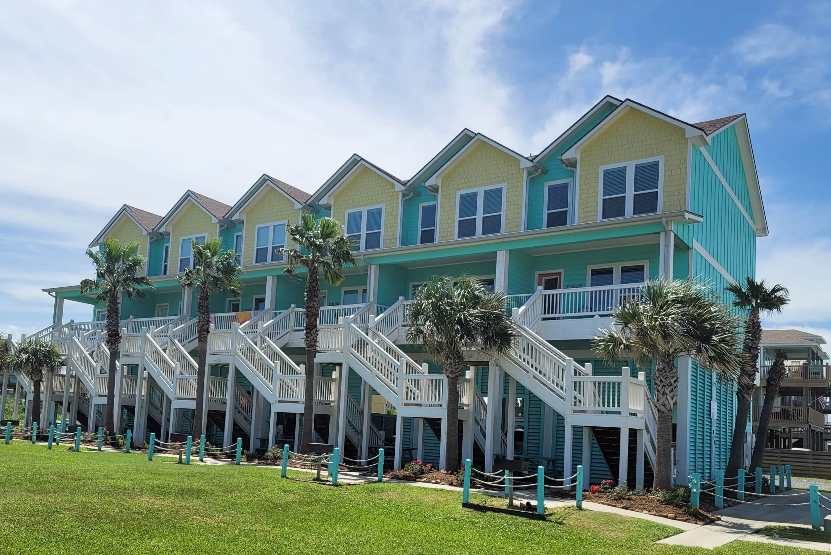 A row of multi-story buildings with balconies and staircases, surrounded by palm trees and a grassy area.