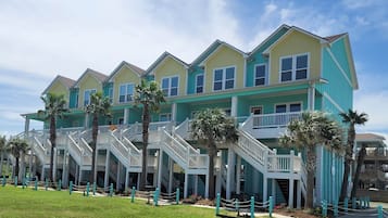 A row of multi-story buildings with balconies and staircases, surrounded by palm trees and a grassy area.