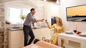 A cozy kitchen with a stone wall, a dining area with a table set for two, and a TV mounted on the wall displaying a sunset scene. - Turrium Heritage Apartment con Patio e Camino Fronte Mare (Kaštel Kambelovac)