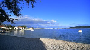 A coastal scene with a pebbly beach, calm water, and a distant view of a town with buildings and a hill. - Turrium Heritage Apartment con Patio e Camino Fronte Mare (Kaštel Kambelovac)