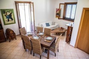 Un coin repas avec une table en bois dressée pour quatre personnes, entourée de chaises en osier. Un canapé blanc et une table basse en bois sont placés contre le mur.