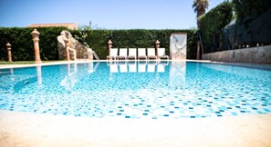 A swimming pool with a tiled floor, surrounded by white lounge chairs and a stone wall.