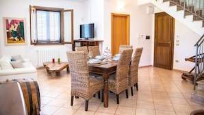 A dining area with wicker chairs, a wooden table set for four, a television on a stand, and a staircase leading to a wooden door. - [✮✮✮✮✮ LUXURY VILLA WITH POOL] - Dimora Fanale (Torre Colonna-Sperone)
