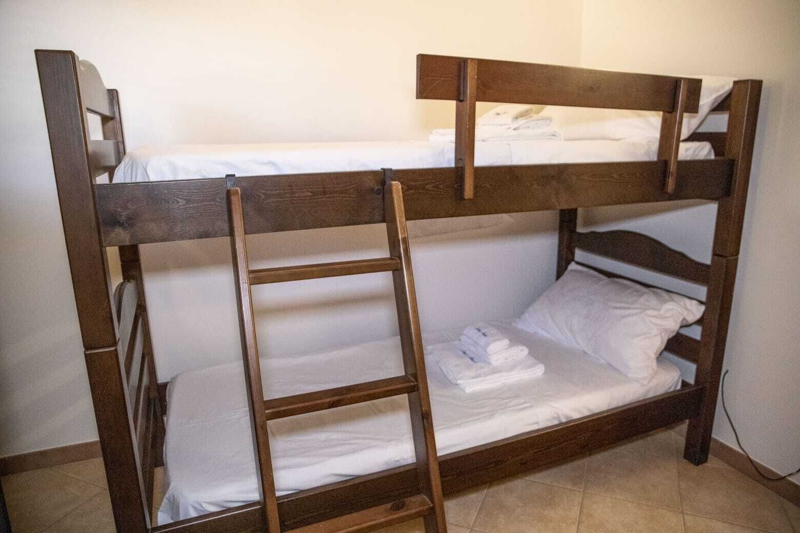 Un llit superposat amb una escala de fusta, roba de llit blanca i una paret sense adorns al fons.