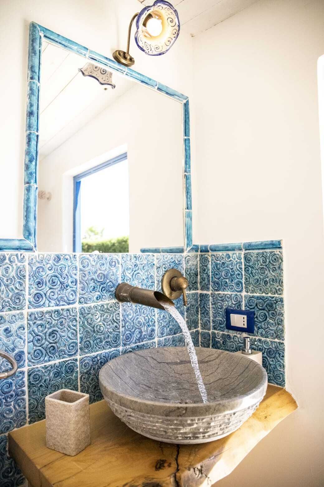 A bathroom with a stone sink, blue patterned tiles, a brass faucet, and a mirror with a decorative frame.