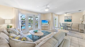 A spacious living room with a large sofa, a coffee table, and a TV. There is a dining area with a table and chairs, and a view of a pool outside. - Impressive Sunsets, Pet-friendly, Heated Pool- Nostra Palazzo on the Canal - Roelens Vacations (Cape Coral)