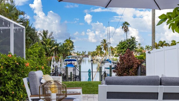 A patio with a white sofa, a wooden table, and a decorative lantern. In the background, there is a marina with boats and palm trees. - Impressive Sunsets, Pet-friendly, Heated Pool- Nostra Palazzo on the Canal - Roelens Vacations (Cape Coral)