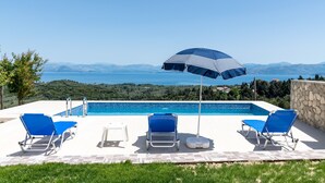 A poolside area with a view of the sea and mountains, featuring blue lounge chairs and a striped umbrella.