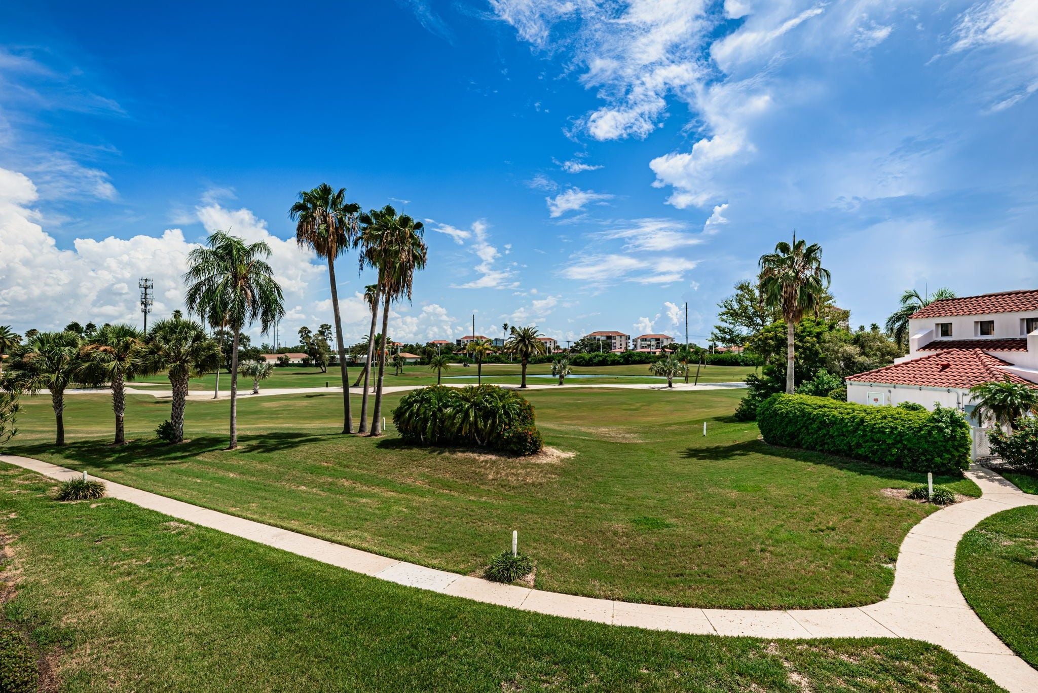 A well-maintained golf course with a clear sky, palm trees, and a pathway leading to a building with a red-tiled roof.