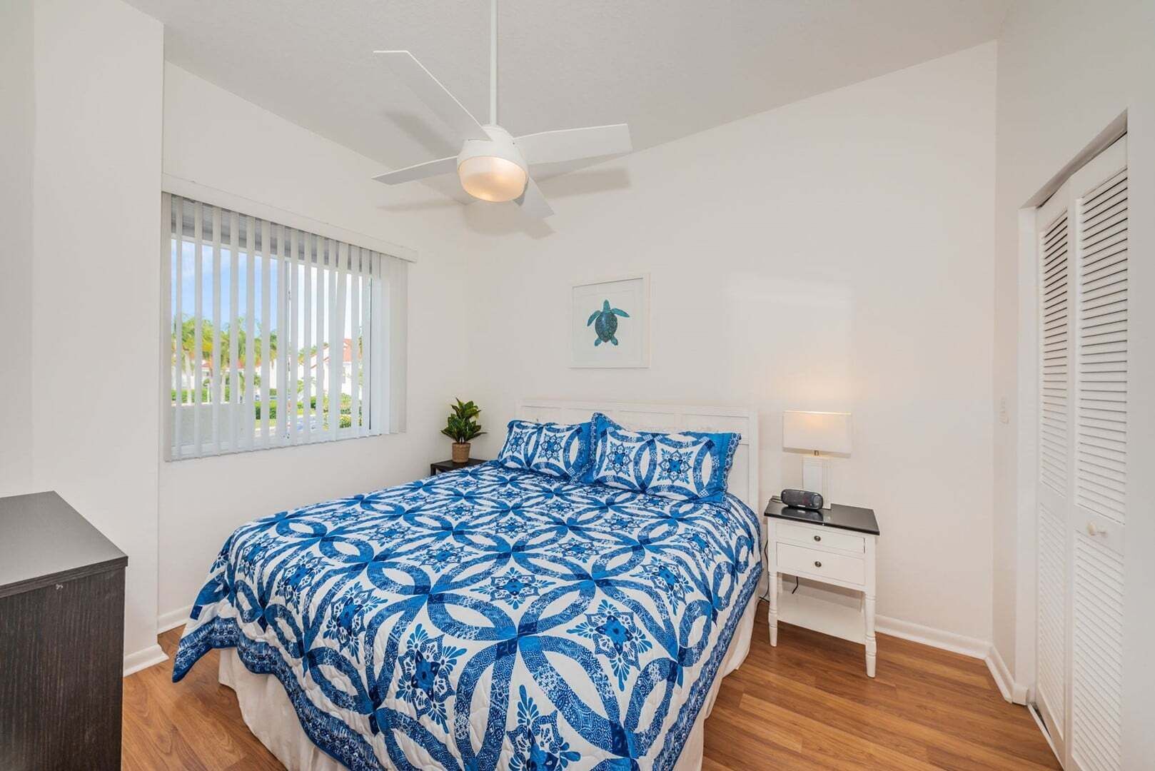 A bedroom with a bed, a nightstand, a ceiling fan, a window with blinds, and a door with louvered shutters.