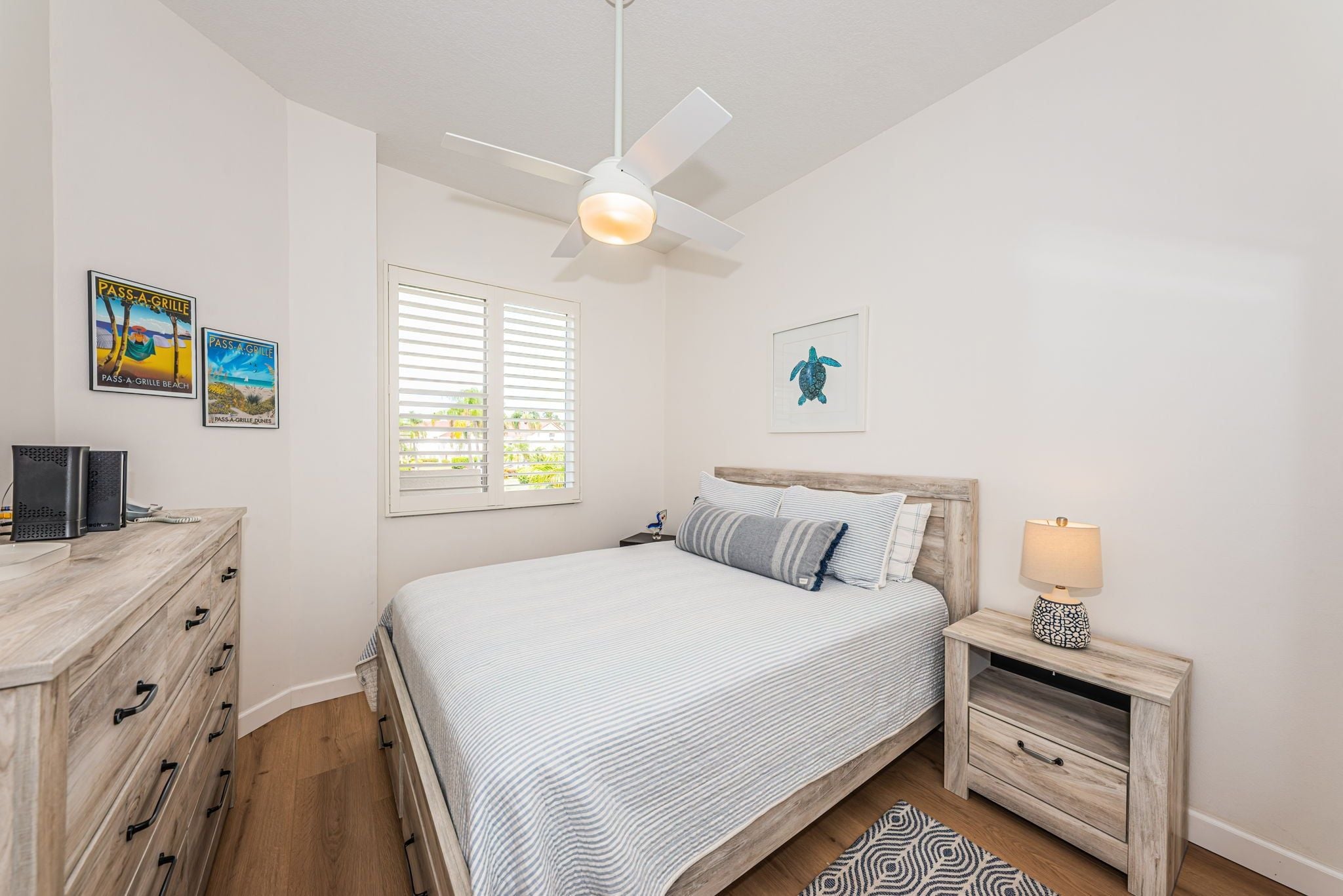 A bedroom with a bed, a nightstand, a desk, a ceiling fan, and two wall-mounted pictures.